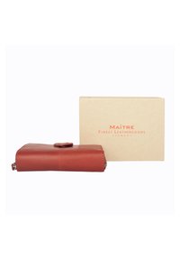 Czerwony skórzany portfel o gładkiej fakturze i zaokrąglonym zapięciu, umieszczony obok beżowego pudełka kartonowego z napisem "MAÎTRE Finest Leathergoods."