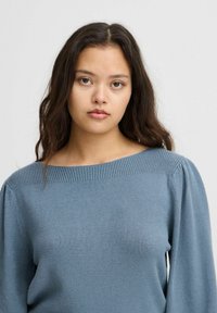 Pull en tricot bleu avec un large col bateau, des détails côtelés sur les épaules et des manches longues bouffantes. Texture douce, design décontracté.