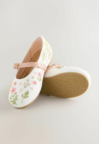 White flat shoes with floral embroidery in pink and green, featuring a pink strap and textured rubber sole with butterfly patterns.