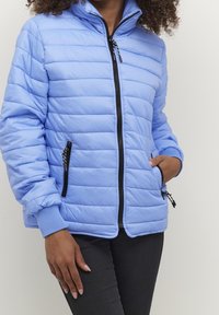 Mujer con pelo rizado que lleva una chaqueta acolchada de color azul claro, pantalones negros, con una mano en el bolsillo y la otra relajada a su lado.