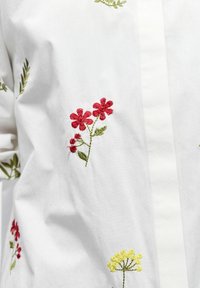 Hvit skjorte i stoff med fargerik blomsterbrodering i rødt, grønt og gult. Designet inkluderer stilker og blader med detaljert søm.