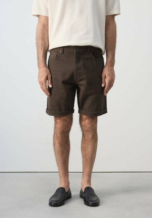 Hombre con pantalones cortos enrollados marrón oscuro, una camiseta sencilla de manga corta color beige claro y zapatos negros sin cordones, de pie en el interior.