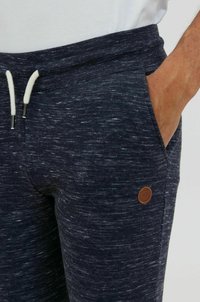 Navy blue joggers with a textured pattern, equipped with an elastic drawstring waist, side pockets, and a small brown leather logo accent.