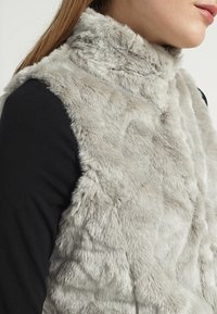 Ljusgrå fuskpälsväst med hög krage och texturerad yta, kombinerad med en svart långärmad topp. Synliga sömdetaljer längs kanterna.