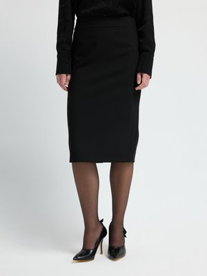 Jupe crayon noire en tissu extensible, présentant une silhouette ajustée. Associée à des chaussures à talons hauts noires et des collants noirs transparents.