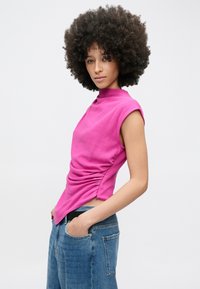 Tânăr cu păr creț în stil afro, purtând un top roz aprins fără mâneci, cu adunături, și blugi albaștri, pozând cu mâinile în buzunare, pe un fundal alb.