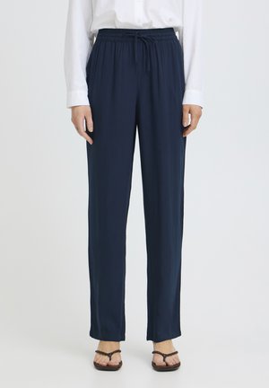 Osoba ubrana w luźne granatowe spodnie ze ściągaczem, białą koszulę z długim rękawem i brązowe plecione sandały, stojąca na tle jednolitego jasnego tła.