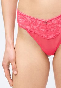 Γυναίκα που φοράει bikini εσώρουχα από κοραλί ροζ δαντέλα και δίχτυ, με ένα μικρό γοητευτικό καρδιά στο κέντρο της μέσης.