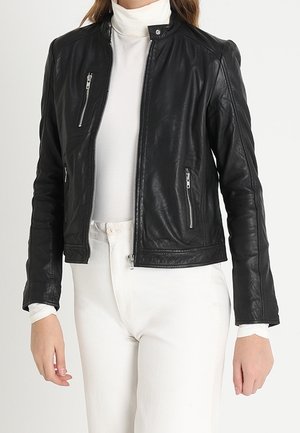 Frau trägt eine schwarze Lederjacke über einem weißen Rollkragenpullover und weißen Hosen, steht mit entspannten Armen an den Seiten.