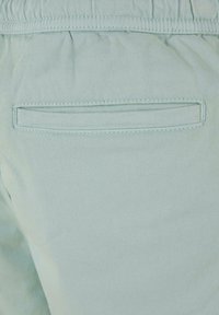 Tissu bleu clair avec une texture lisse, comportant une poche arrière avec une ouverture rectangulaire et des coutures le long des bords. Ceinture élastique visible.
