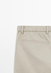 Beżowe bawełniane spodnie z elastycznym paskiem w talii, z pętelkami na pasek i jedną tylną kieszenią. Gładka tekstura i minimalistyczny design.