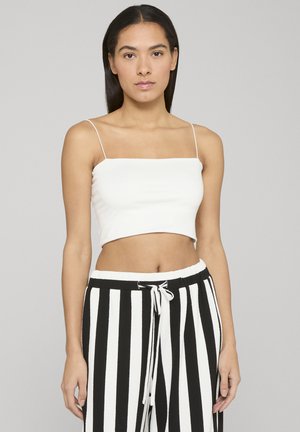 Ung kvinde med langt mørkt hår iført en hvid crop top med spaghetti stropper og sort-hvide bukser med lodrette striber og snøre i taljen.