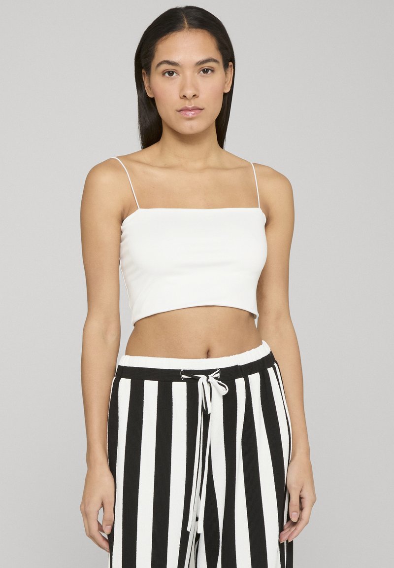 Młoda kobieta z długimi ciemnymi włosami, ubrana w biały top na cienkich ramiączkach oraz czarno-białe pionowo paskowane spodnie z wiązanym paskiem.