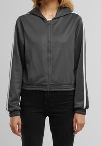 Gray cropped jacket with hood, zip front, and elastic cuffs. Features white striped accents along sleeves and smooth, lightweight fabric.