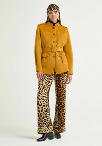 Giacca giallo senape con cintura e spalline sopra un dolcevita nero, abbinata a pantaloni larghi con stampa leopardata marrone e stivaletti neri.