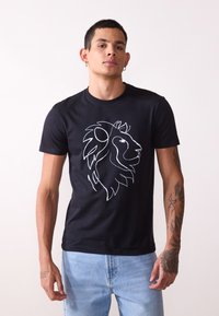 Zwarte katoenen t-shirt met een witte omtrek van een leeuwenkopontwerp aan de voorkant. Model draagt lichtblauwe jeans.
