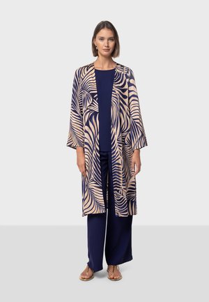 Printed long kimono in beige and navy, featuring a leaf pattern. Paired with a navy top and wide-leg pants; sandals complete the look.