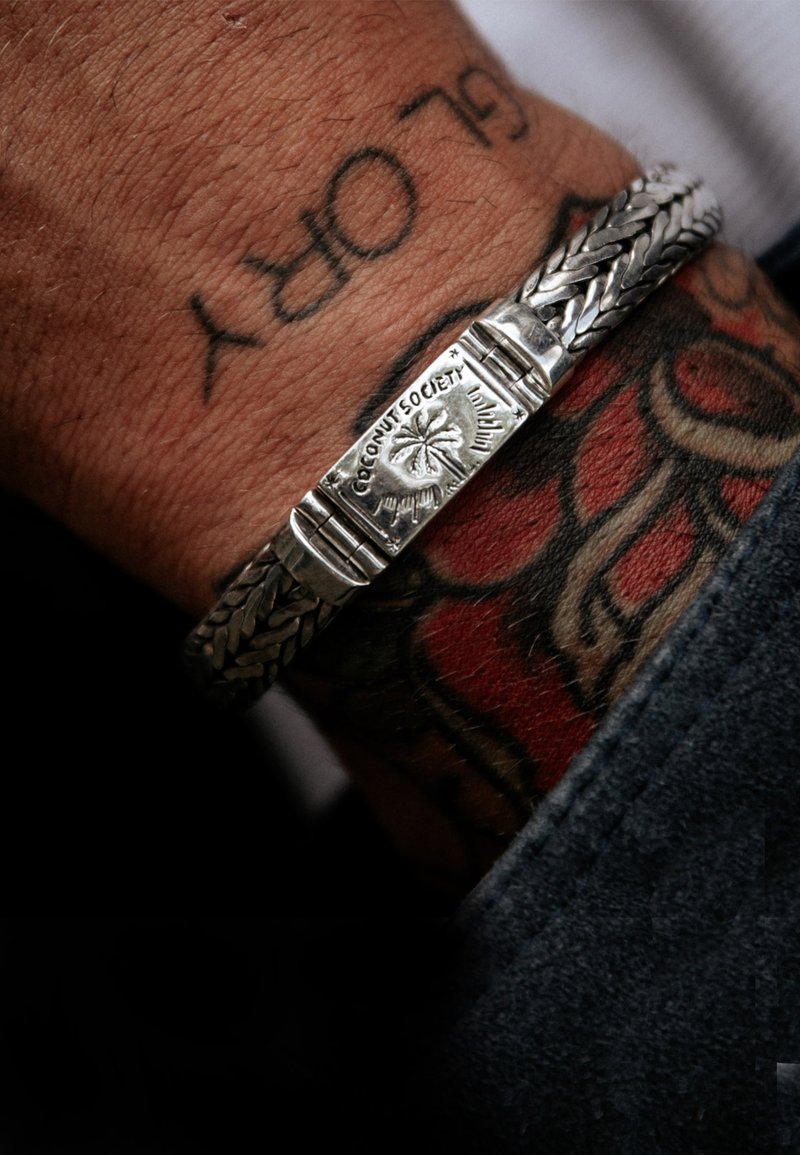 Silverflätad armband med ett platt, graverat lås som har en logotyp och en blommönster. Hud med tatueringar syns under armbandet.