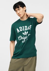 Νεαρός άνδρας με μαύρα μαλλιά, που φοράει σκούρο πράσινο T-shirt Adidas Originals και χακί σορτς, ποζάρει με το χέρι πίσω από το λαιμό.