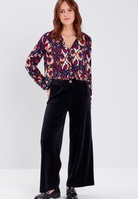 Blouse à manches longues à motifs colorés avec un col en V, associée à un pantalon large en velours côtelé noir avec un détail de bouton à l'avant.