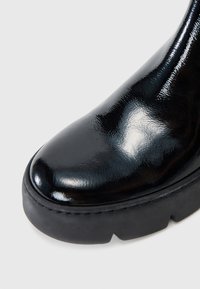 Botte de cheville en cuir verni noir avec un bout rond, avec une semelle épaisse noire et un minimum de détails de couture. Texture de surface lisse.