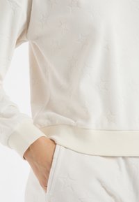 Biała bluza z wyraźnym wzorem gwiazdy, wykonana z miękkiego polaru, z prążkowanymi mankietami i dołem oraz detalem z ręką w kieszeni.