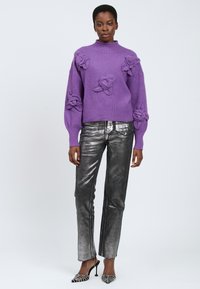 Frau trägt einen lila Pullover mit 3D-Blütendetails, metallisch silberne Hose und schwarze mit Nieten besetzte High Heels vor einem grauen Hintergrund.