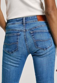 Pantalones vaqueros de mezclilla azul de corte ajustado, con costuras en contraste, dos bolsillos traseros y una etiqueta de cuero marrón en la cinturilla.