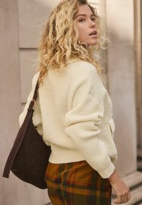 Ivory, textured sweater with ribbed sleeves, paired with plaid, corduroy pants featuring warm tones of orange and green; brown woven bag.