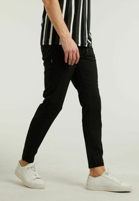Zwarte slim-fit broek met een gladde textuur, voorzien van zijnaad en gedragen met witte sneakers. Model poseert halverwege een stap.