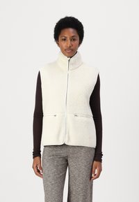 Kraftig fleeceväst med hög krage, framskjutande dragkedja, två dragkedjefickor och mjuk textur, bärs över en mörkbrun långärmad topp.