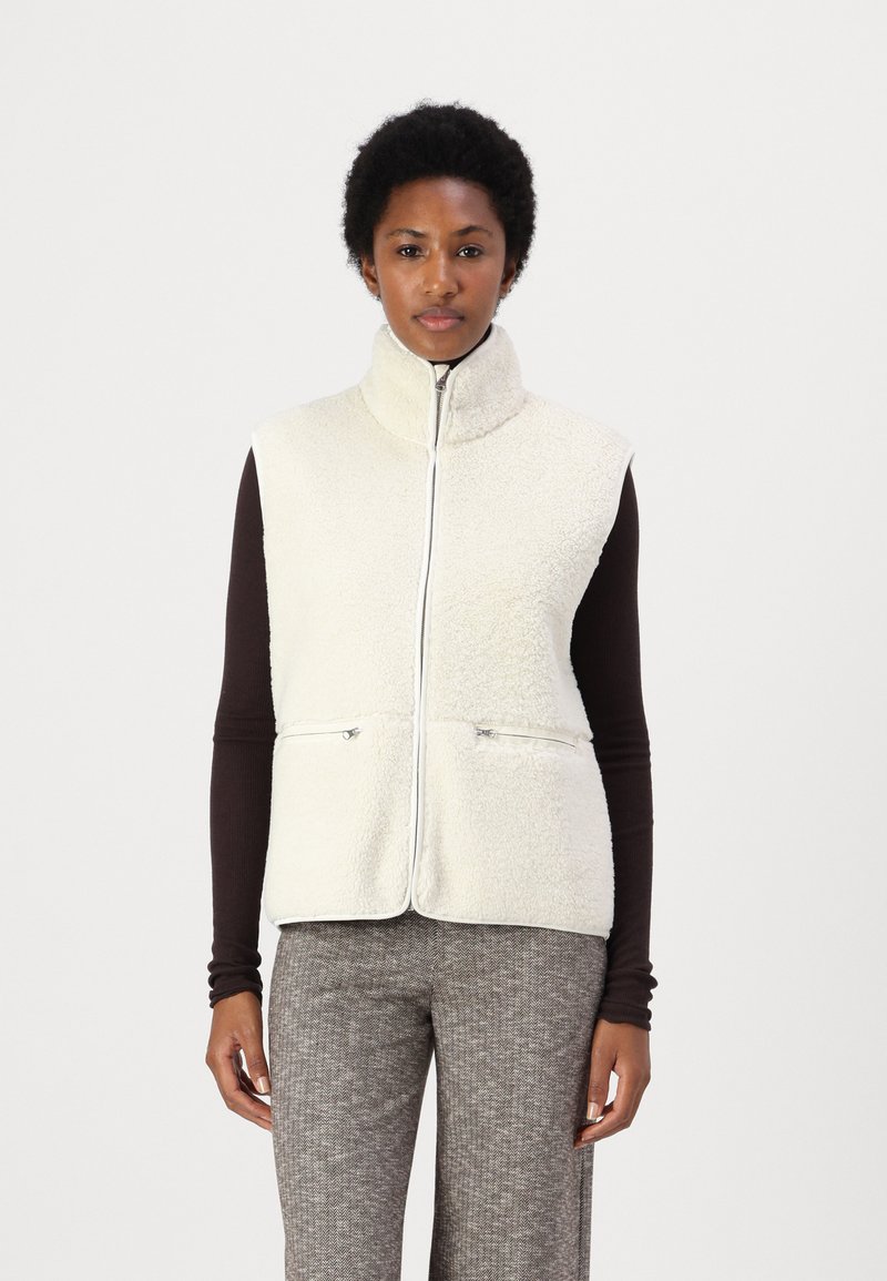 Kraftig fleeceväst med hög krage, framskjutande dragkedja, två dragkedjefickor och mjuk textur, bärs över en mörkbrun långärmad topp.