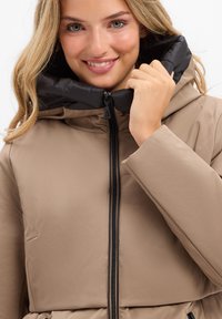 Tan pufferjack met een zwarte, gequiltde capuchon, centrale ritssluiting aan de voorkant en een gladde textuur. Heeft een getailleerde pasvorm en minimale details.
