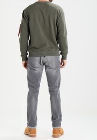 Uomo in piedi di spalle, indossando una felpa verde oliva, jeans grigi e stivali da lavoro beige su sfondo bianco.