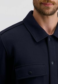 Navyblauwe jas met een knoopsluiting aan de voorkant, een opklapbare kraag en een borstzak met klep. Gemaakt van een gladde, gestructureerde stof.