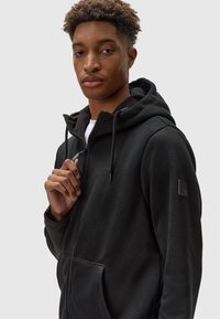 Felpa nera con zip, caratterizzata da un tessuto testurizzato, cappuccio con cordino regolabile e tasche frontali, con un patch logo sulla manica sinistra.
