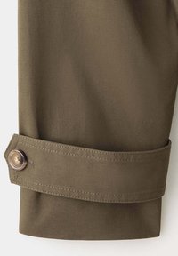 Gros plan d'une manche de manteau marron avec une bride cousue maintenue par un bouton marron près du poignet.