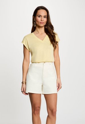 Donna che indossa una maglietta a maniche corte color giallo pallido con scollo a V e pantaloncini bianchi a vita alta, accessoriata con orecchini e braccialetti dorati.