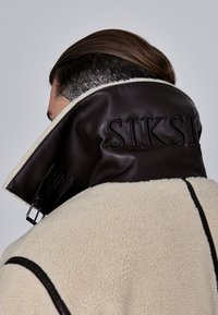 Cream shearling coat with a high brown leather collar featuring embossed "SIKSI" text, finished with black accent stitching.