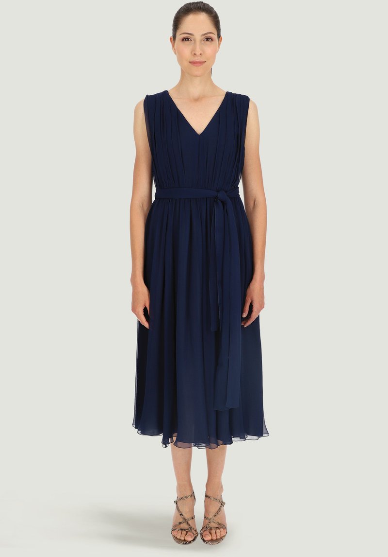 Granatowa, do kolan sukienka z dekoltem w kształcie litery V, plisowanym gorsetem i tkaninowym paskiem. Przezroczysty brzeg i miękka struktura, w połączeniu z sandałami na cienkich paskach.