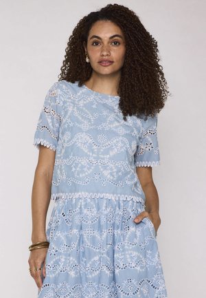 Donna con capelli ricci che indossa una maglietta azzurra ricamata a maniche corte e una gonna abbinata, in piedi con la mano in tasca su sfondo semplice.