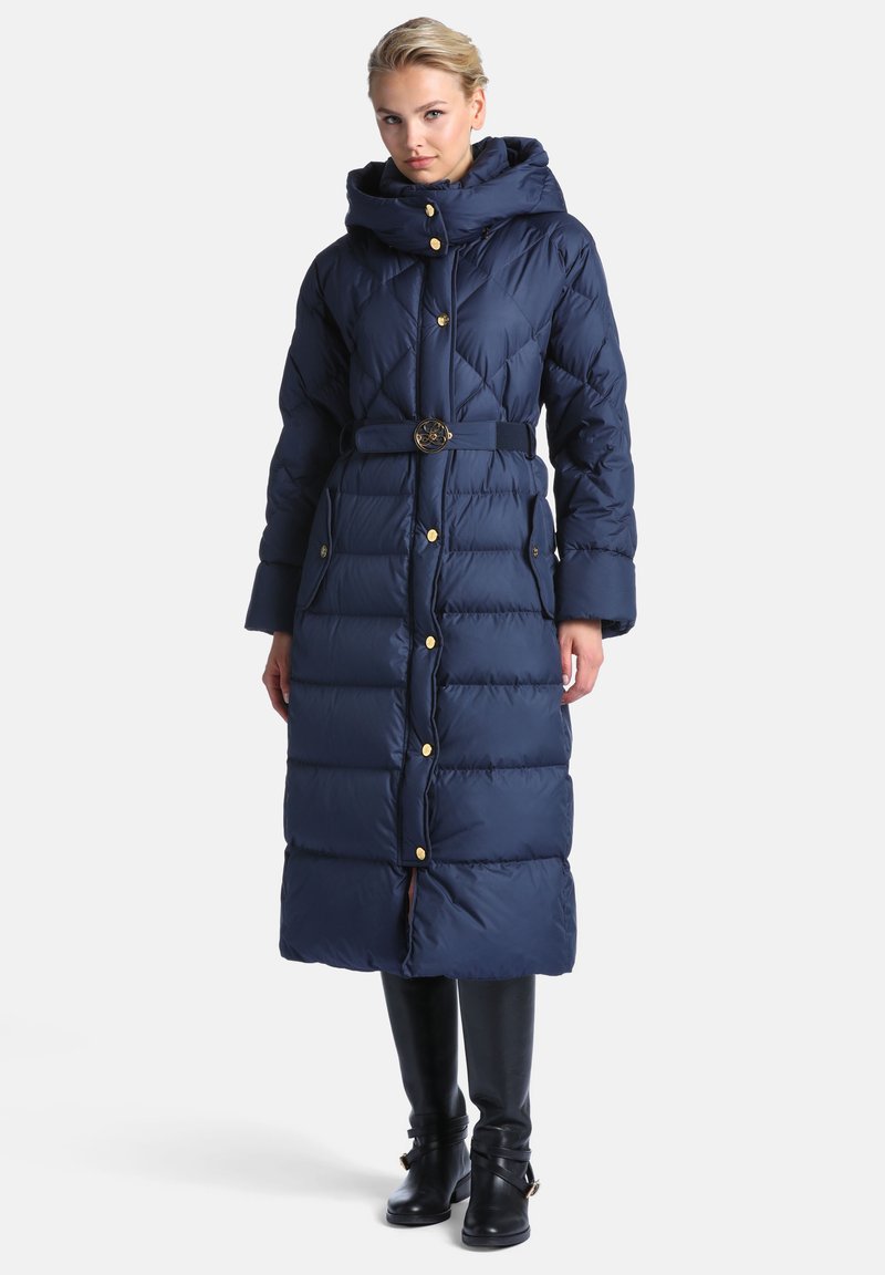 Femme portant un long manteau matelassé bleu marine avec une capuche, taille ceinturée, boutons dorés, poches latérales et bottes noires montant jusqu'aux genoux.