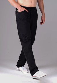 Schwarze, gerade geschnittene Hosen mit glatter Textur, die über Fronttaschen, einen Knopfverschluss und subtile Stickdetails verfügen. Getragen mit weißen Turnschuhen.