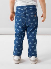 Pantalones de mezclilla azul con patrones de corazones azul claro, cinco bolsillos y un corte relajado. El niño lleva zapatillas blancas con acentos grises.