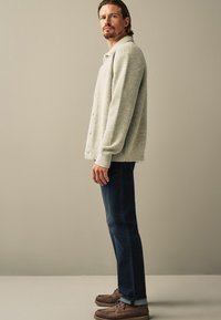 Lichtgrijs geribbeld gebreid shirt met knopen, gecombineerd met donkerblauwe jeans en bruin lederen botenschoenen, vanuit de zijaanzicht tegen een neutrale achtergrond.