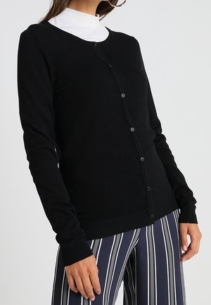 Kvinde iført en sort knaplukket cardigan over en hvid top med høj hals og marineblå bukser med hvide lodrette striber.