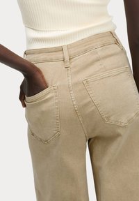Béžové kalhoty s vysokým pasem a rovnými nohavicemi, s zadními kapsami a strukturovanou tkaninou. Viditelné švy podél švů.