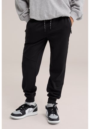 Persoon die zwarte joggingbroek met trekkoorden draagt, witte en zwarte sneakers, en een grijze trui met handen in de zakken.
