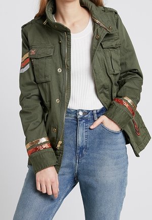 Personne portant une veste vert olive avec des rayures de sequins rouges et dorées sur les manches, un haut blanc côtelé et un jean bleu taille haute, main dans la poche.