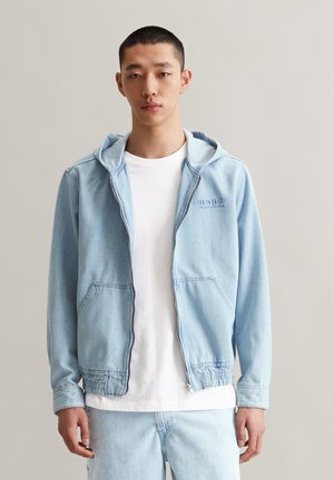 Lichtblauwe spijkerjack met een capuchon, ritssluiting en twee zijzakken. Heeft elastische manchetten en een casual pasvorm, gelaagd over een wit t-shirt.